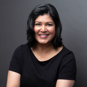 Headshot of a south Asian woman with dark shoulder length hair with a black t-shirt on a gray background